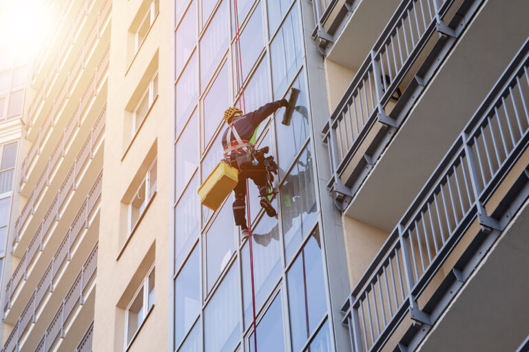 Ein Gebäudereiniger hängt an Seilen und wäscht die Fenster einer hohen Glasfassade.