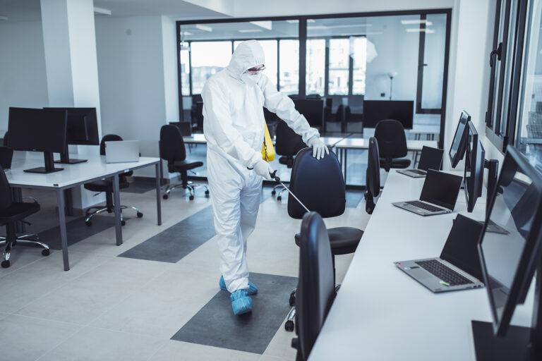 Mitarbeiter in Schutzanzug desinfiziert Stühle und Arbeitsplätze im Büro.