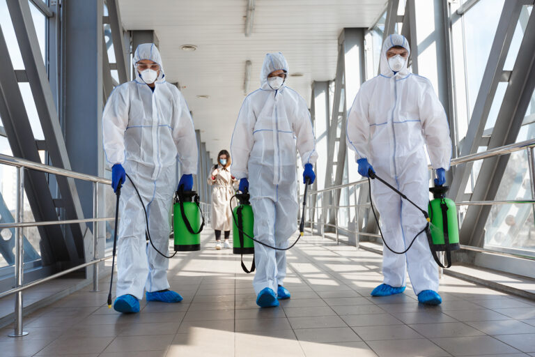 Desinfektionsteam in Schutzanzügen läuft durch ein Gebäude und sprüht Mittel.