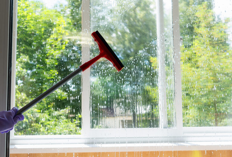 Hand mit Gummihandschuh zieht mit rotem Abzieher Seifenwasser von einer verglasten Fensterfläche; draußen sind grüne Bäume sichtbar.