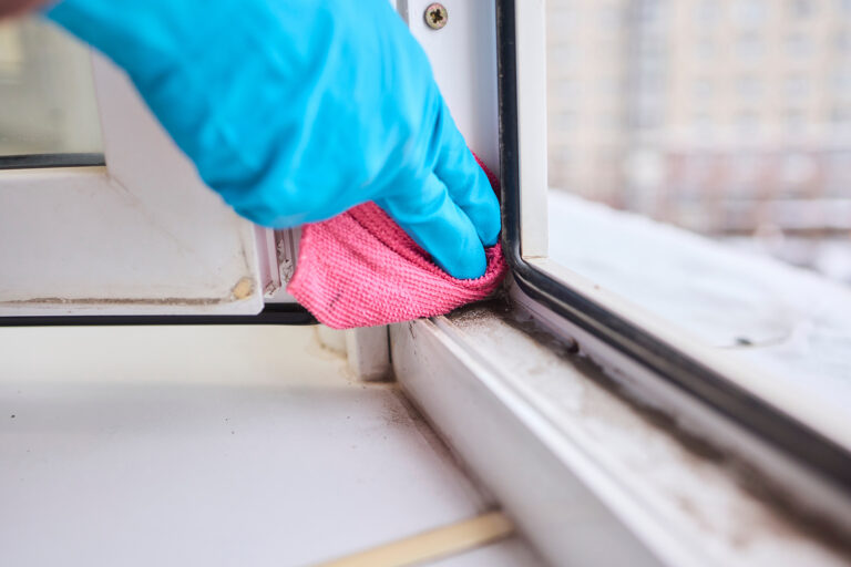 Hand in blauem Handschuh reinigt mit pinkem Mikrofasertuch die verschmutzte Fensterschiene.