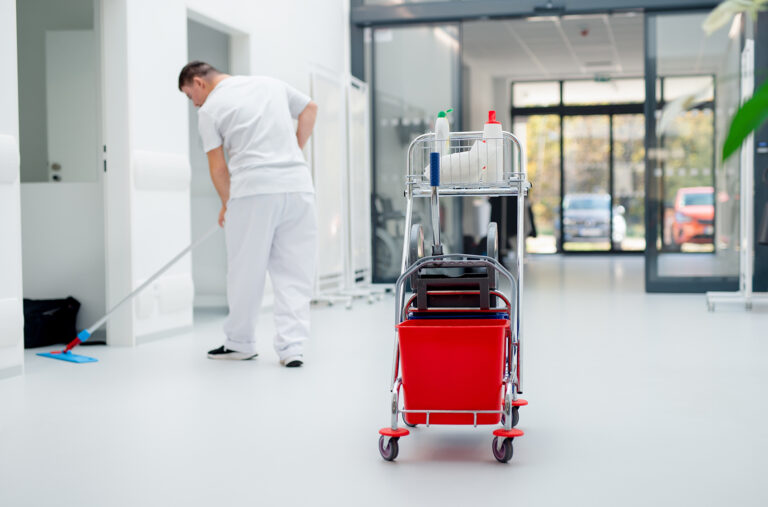Reinigungswagen mit rotem Eimer in Klinik-Korridor.