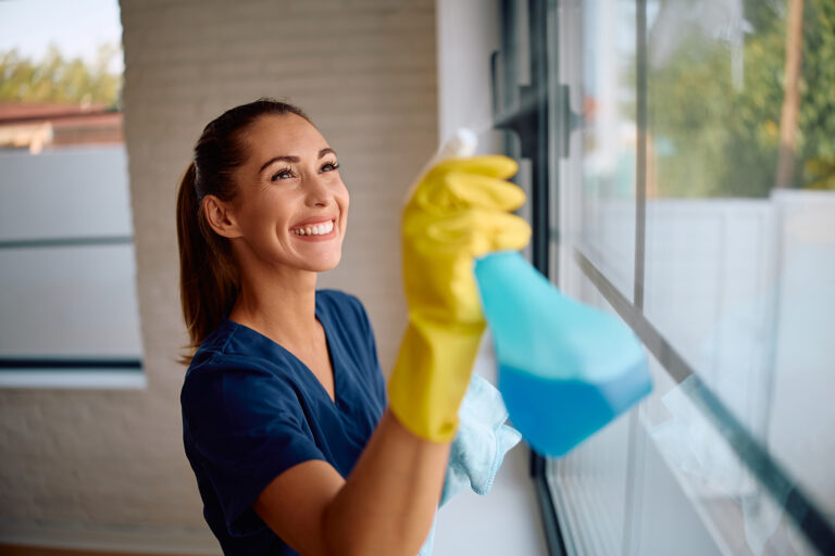 Lächelnde Reinigungskraft sprüht Fensterreiniger auf eine Glasscheibe und wischt mit Tuch; gelbe Gummihandschuhe und blaue Arbeitskleidung.